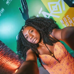 A woman with curly hair wearing a patterned orange dress takes a selfie in front of a colorful wall with geometric designs and text in Portuguese. The background includes green, yellow, and brick elements.