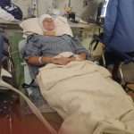 A person lies in a hospital bed with a bandaged head, wearing a hospital gown and covered with a blanket. Medical equipment and monitors are visible in the background, and another person stands nearby.