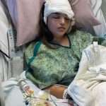 A young girl with a bandaged head lies in a hospital bed, eyes closed. She wears a hospital gown and has an IV in her arm, connected to tubing and a bag of fluid with red liquid.
