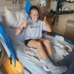 A woman sitting up in a hospital bed smiles and gives two thumbs up. She wears a gray t-shirt, black shorts, and gray grip socks. Medical equipment and a monitor are visible in the background.
