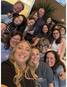 A group of fourteen adults, smiling and posing closely together for a selfie indoors. Most are women, and the atmosphere appears cheerful and friendly, with some people wearing glasses and casual clothing.