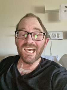 A man with glasses and a beard smiles while sitting in a hospital room. He has medical tape on his upper chest and is wearing a dark shirt. Medical equipment and a partial sign are visible in the background.