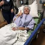 A woman in a hospital bed wearing a gown and hair cap gives a thumbs up and smiles. Medical equipment and a staff member are visible in the background.