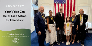 Six people, including a child, stand smiling in front of an American flag at an advocacy event for Ellies Law, organized by The Bee Foundation for Brain Aneurysm Prevention. A sign encourages taking action for the cause.
