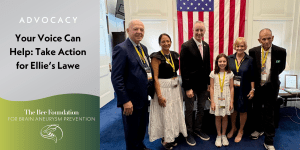 Six people, including a young girl, stand smiling in front of a large American flag in a formal room. A banner on the left reads, Your Voice Can Help: Take Action for Ellies Law—The Bee Foundation for Brain Aneurysm Prevention..