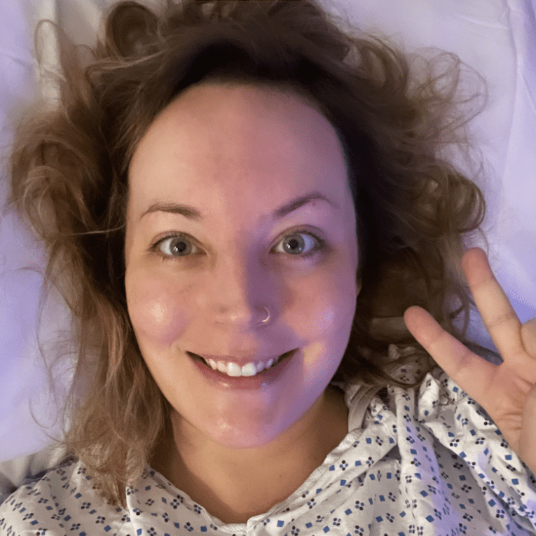 A woman with wavy hair lies on a bed, wearing a hospital gown. She smiles at the camera and makes a peace sign with her hand.