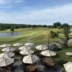 Outdoor patio with tables and white umbrellas overlooks a lush golf course, winding cart paths, trees, a small pond, and a partly cloudy blue sky in the background.