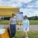 Two women in hats stand and smile under a yellow tent at an outdoor event. One holds a sign for The Tee Store. A table with items and trees in the background are visible under a partly cloudy sky.