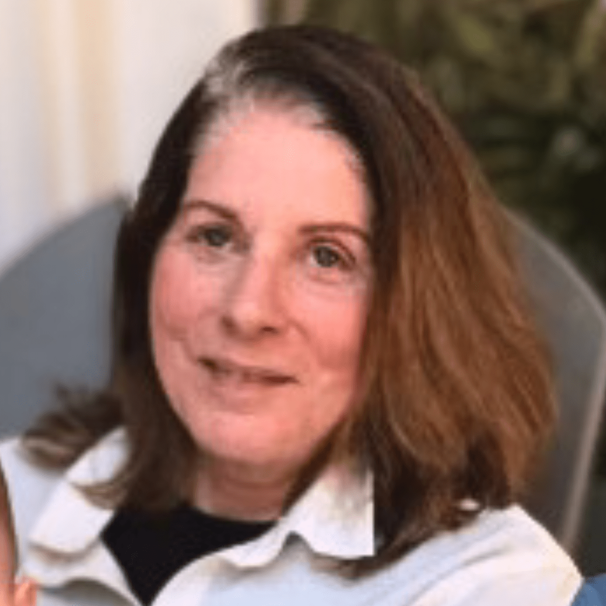 A woman with shoulder-length brown hair, wearing a white collared shirt and black top, sits outdoors in a chair, smiling softly. There is greenery and a white curtain in the background.