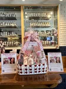 A white gift basket with heart decorations, filled with pink and white items, sits on a table. Two flyers promoting a raffle surround it. In the background, a display case holds many collectible figurines.