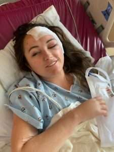 A woman in a hospital bed wears a hospital gown, with a bandage on her forehead and one eye swollen shut. She is holding a white bag, smiling slightly, and appears to be recovering from a head injury.