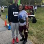 Two people stand smiling at a race finish line, one holding a bell and wearing a race bib numbered 8869. A digital race timer behind them reads 9:24. Trees and other participants are in the background.