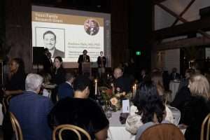 People seated at tables during a formal gala; a speaker stands at a podium with a projected slide showing the Yezzi Family Research Grant and a photo of Andrew Hale, MD, PhD. Elegant decor and candles are visible throughout the room.