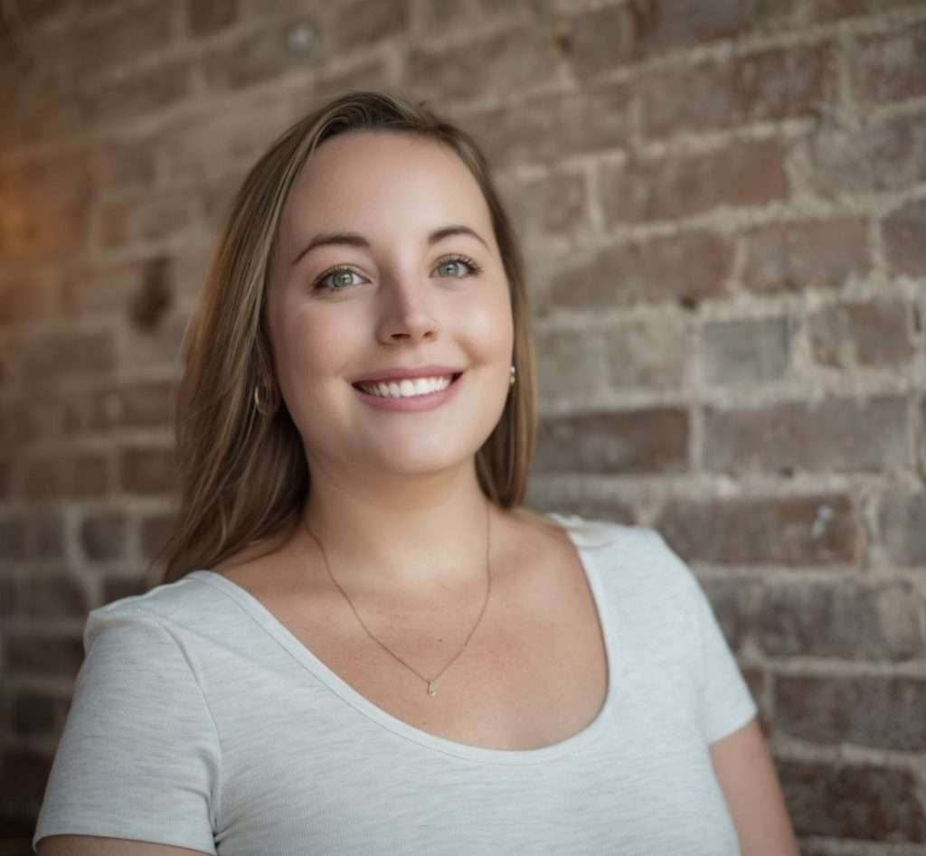 A woman with long light brown hair, wearing a white shirt and a delicate necklace, smiles at the camera. She stands in front of a brick wall background.
