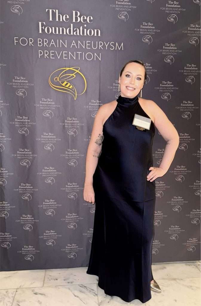 A woman in a sleeveless black gown stands and smiles in front of a backdrop for The Bee Foundation for Brain Aneurysm Prevention.