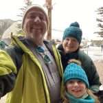 A man and two smiling children, all wearing winter jackets and knit hats, take a selfie outdoors in a snowy area with trees, people, and buildings in the background.