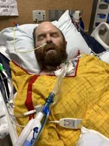 A bearded man lies unconscious in a hospital bed with a yellow blanket, connected to medical tubes and equipment, including a ventilator and a nasal feeding tube. Hospital monitors and devices are visible in the background.