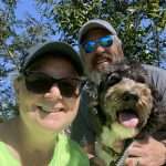 A woman and man wearing sunglasses and hats smile outdoors with a happy black and white dog, all enjoying a sunny day under green trees.