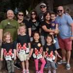 A group of four adults and five children pose together outdoors. The children wear matching Minnie Mouse shirts, and one has a butterfly face paint. The adults are smiling, and two hold children in their arms. Rocks and plants are in the background.