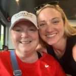 Two smiling women pose for a selfie indoors; one wears a white cap and red shirt with a blue strap, the other wears a sleeveless black top and sunglasses on her head. Both appear happy and close together.