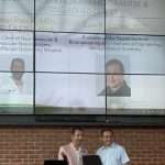 Two men stand and smile while one hands a certificate to the other. Behind them, a large screen displays photos and names of award recipients and guests, including Dr. Tudor G. Jovin and Dr. Juan R. Cebral.