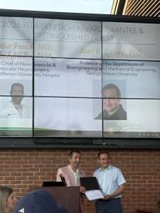 Two men stand and smile while one hands a certificate to the other. Behind them, a large screen displays photos and names of award recipients and guests, including Dr. Tudor G. Jovin and Dr. Juan R. Cebral.