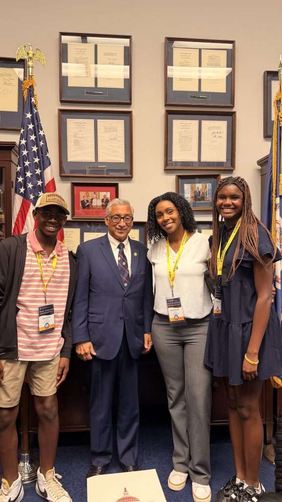 Three young people stand with an older man in a suit in an office with framed documents on the wall and American flags. All are smiling and wearing event badges.