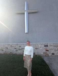 A woman stands on grass in front of a large cross mounted on a stone and concrete building wall, with sunlight streaming from the upper left corner.
