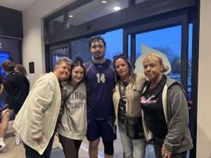 A group of five people pose and smile together indoors near a glass door. One person in the center wears a purple sports uniform with the number 14, while the others wear casual clothes.