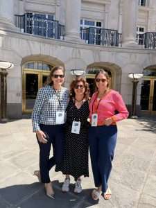 Three smiling women stand together outside a stone building with large windows and glass doors, each wearing conference badges around their necks. They are dressed in business-casual clothing and standing on a stone walkway.