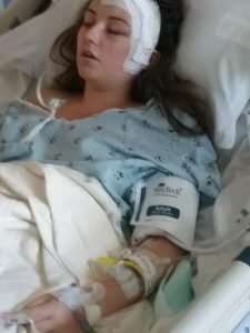 A young woman lies in a hospital bed wearing a hospital gown, with medical monitors and bandages on her head and wrist, and a blood pressure cuff on her arm. She appears to be sleeping or unconscious.