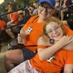 A woman and a smiling girl wearing orange Houston Astros shirts sit together at a stadium. The woman holds a drink and has her arm around the girl, who is wearing glasses. Fans in orange shirts fill the seats behind them.