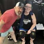A man in a red shirt and cap leans toward a smiling woman seated on a bench, wearing a CrossFit Open 2022 tank top, knee sleeves, and glasses. Chalkboard walls and gym equipment are visible in the background.
