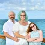 A smiling family of three poses by a wooden railing with the ocean in the background. The man and woman wear white, and the young girl with red hair wears sunglasses and a blue dress. The sky is partly cloudy.