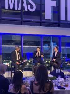 Three men in suits sit on stage with microphones during a panel discussion at a formal event. They face an audience with dinner tables, and large windows reveal city lights outside.
