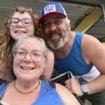 A smiling woman, man, and young girl pose together for a selfie in a garage. The woman and girl wear glasses; the man wears a blue cap and tank top. Bicycles are visible in the background.