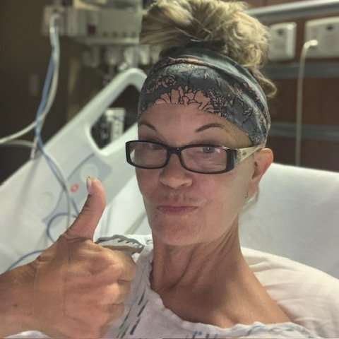 A woman wearing glasses and a headband gives a thumbs up while lying in a hospital bed, smiling confidently. Medical equipment and monitors are visible in the background.
