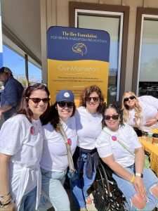 Five women, all wearing white shirts, smile and pose together at an outdoor event in front of a banner for The Bee Foundation for Brain Aneurysm Prevention. One woman is in a wheelchair; they appear cheerful and united.
