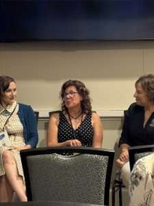 Three women sit and talk together in a conference room, with two upholstered chairs visible in the foreground. One woman speaks while the others listen attentively. They appear to be engaged in a panel discussion.