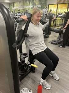 A woman in workout clothes uses a chest press machine at the gym, appearing to exert effort. Cardio equipment and motivational wall art are visible in the background.