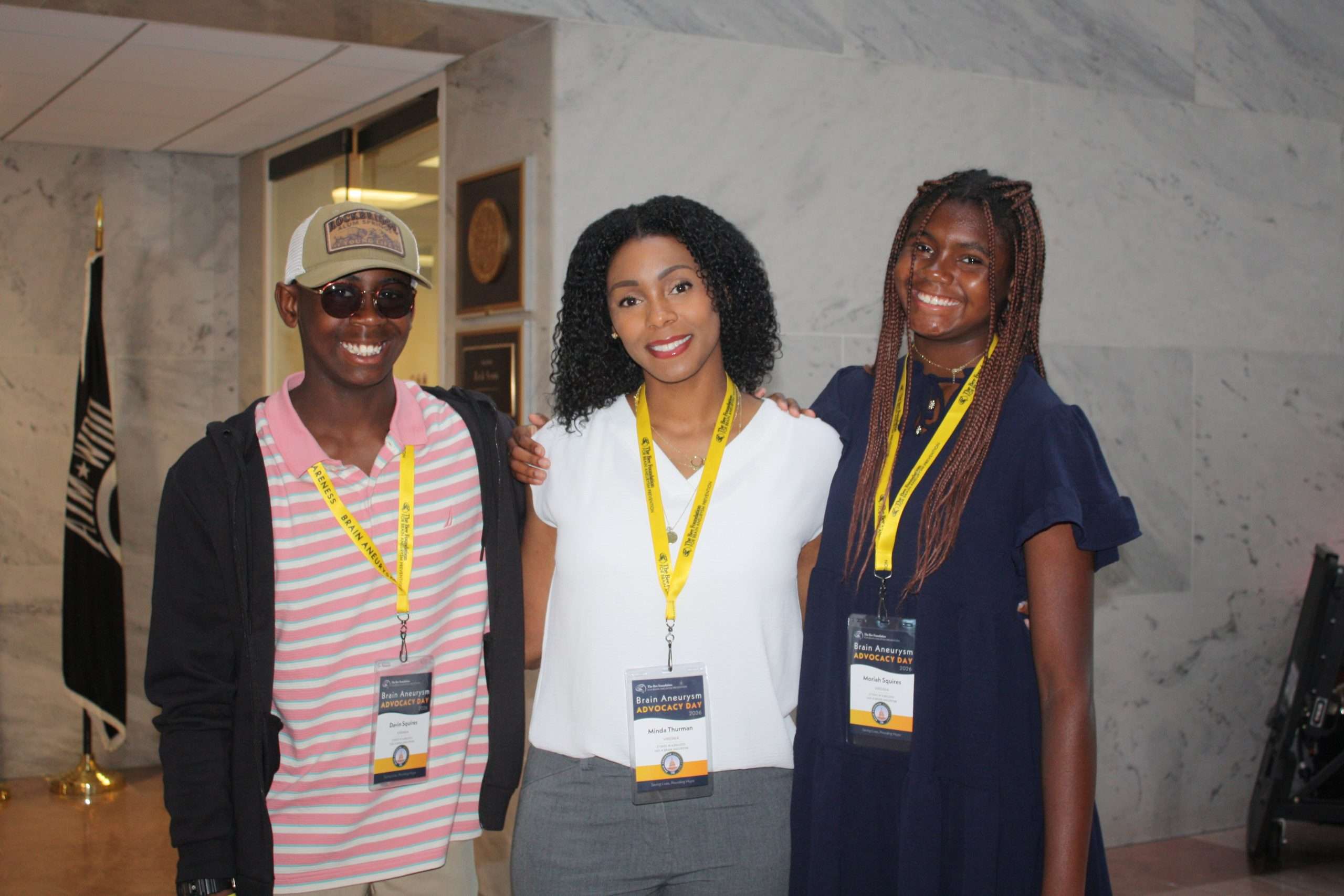 Three people stand indoors smiling at the camera, all wearing conference badges with yellow lanyards. The person in the middle has curly hair and a white shirt; the others are on either side, one in a striped shirt and hat, the other in a navy dress.
