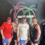 Four people pose and smile in front of a mural with palm trees and the words “CrossFit Anayah.” Three wear matching American flag shorts, and two wear “CrossFit Anayah” shirts. The group stands in a gym-like setting.