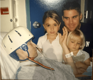 A person in a hospital bed wearing a protective helmet looks at two young girls and a man, who are sitting beside them. The atmosphere appears emotional and supportive.