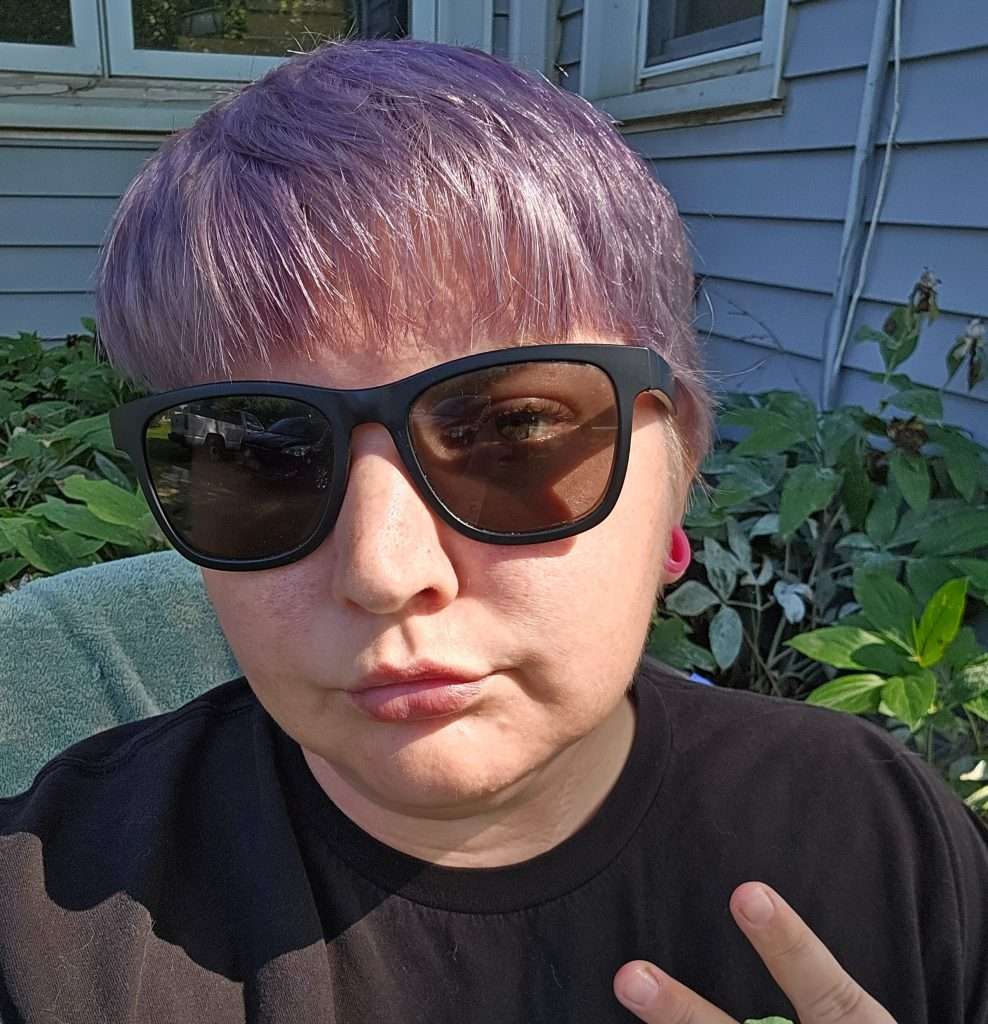 Person with short purple hair and black sunglasses sits outside in front of green plants, making a peace sign with their fingers. They wear a black shirt and have a neutral expression.