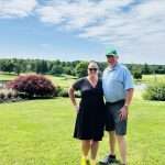 A smiling couple stands on a bright green lawn with a golf course, trees, and a blue sky with scattered clouds in the background. The woman wears a black dress and yellow shoes; the man wears a green cap, patterned polo, shorts, and sneakers.
