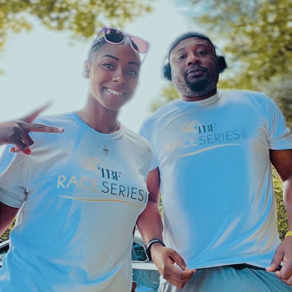 Two people outdoors wearing matching white TBF Race Series shirts. The woman is smiling, wearing sunglasses on her head, and making a peace sign. The man is wearing headphones and looking at the camera. Trees and sunlight are in the background.