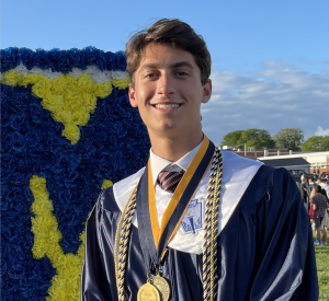 A young man in a graduation cap and gown stands outside, smiling, wearing honor cords and medals. A large blue and yellow floral letter M is visible in the background, with a crowd and buildings behind him.