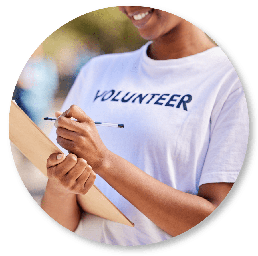 A person wearing a VOLUNTEER t-shirt smiles while holding a clipboard and pen, appearing to write or take notes outdoors.