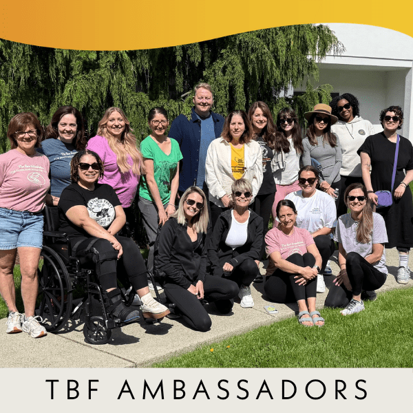 A group of sixteen people, diverse in age and appearance, pose together outside on a sunny day. Some are sitting, one uses a wheelchair, and all are smiling. Text at the bottom reads TBF AMBASSADORS.