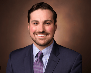 A man in a navy suit, light blue shirt, and purple tie smiles at the camera against a brown, softly blurred background.